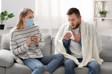 Woman in protective mask looking at her coughing husband