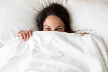 Top view close up playful pretty girl hiding face, peeking from blanket, lying on soft pillow in bed, looking at camera, beautiful young woman shy, covering face with duvet, having fun at home