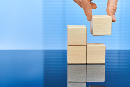 Four wooden cubes on a blue background in two rows with a reflection on the surface. A man's hand raises the upper cube on the left. Free space for letters, numbers, symbols, or labels.