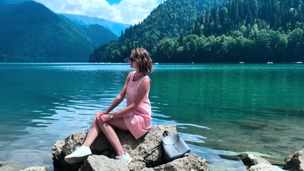 A girl in a pink dress and sunglasses sits on the Bank of a blue lake on a large rock on a clear Sunny day. Ritsa lake