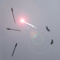 A collection of beauty treatment tools floating in mid air including lash tweezers, mascara wands or eyebrow brushes and strip lash extensions against a plain grey background with a lens flare 