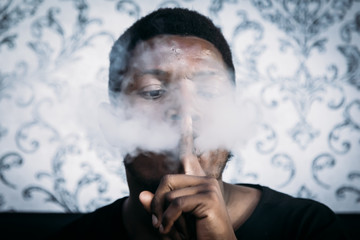 A black man is smoking from his mouth while covering it with a finger