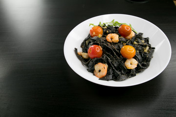 Black Spaghetti or tagliatelle with shrimps, cherry tomato and basil served in a white plate on a dark wooden table. Italian cuisine