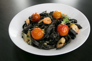 Black Spaghetti or tagliatelle with shrimps, cherry tomato and basil served in a white plate on a dark wooden table. Italian cuisine