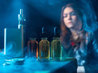 Vape devices are on the table. The oil next to the electronic cigarette. Bottle vaping liquids. A viper girl in the background. Woman in the smoke from vaping. Concept - vape oils of different tastes