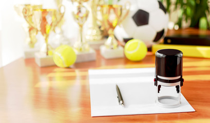 Office workplace with a blank sheet, pen, stamp, soccer and tennis balls, trophies on a blurred...