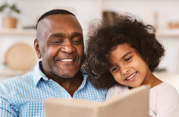Family lockdown hobbies. African American child listening to her grandfather read bedtime story at...