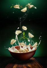 Hot pierogi flying out of the clay bowl with cream and parsley. Some vareniki stay inside the plate.  Green background.