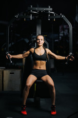 Fototapeta na wymiar Beautiful fitness female with perfect athletic body wearing black sportswear using training simulator for pumping back muscles during sport workout training in modern dark gym.