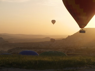 Golden Balloon Rise.Hot Air Balloons with Basket full of People, in Distance and rising out of Meadow. Soft warm colours in sunrise over mystic misty mountaineous landscape.
