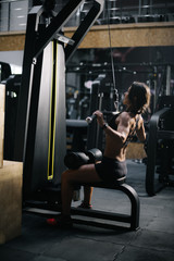 Fototapeta na wymiar Strong unrecognizable sporty woman using training simulator for pumping back muscles in gym, back of view. Concept of professional training in a modern dark gym.