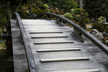 Fototapeta premium Simple wooden walkway across a small stream in a Japanese public park. Local bridge with small planks for slippery protection.