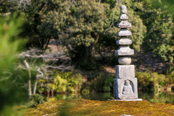 Fototapeta na wymiar Single Japanese Buddhist stupa made of local stones stands on small islet. Oriental stupa is in the middle of a pond surrounded by natural forest. Symbol of peace and calm for East Asia people