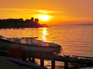 Resting Boat.Covered tiny Boat on in Foreground, golden glowing Sky and Sea in Sunset at Izmit Bay. Silhouette of Cape with Trees and Houses, Osman Gazi Bridge in the Far.