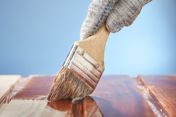 Man's hand with a paint brush painting wooden surface on a blue background.