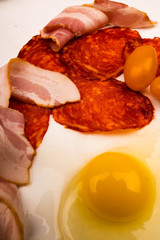 A broken chicken egg without a shell, slices of sausage and bacon and tomatoes on a white background. Close up.