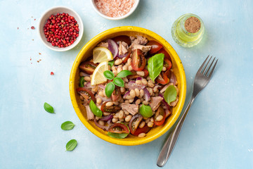 Healthy salad with tuna, white beans, cherry tomatoes, red onion and basil leaves in ceramic bowl on concrete background