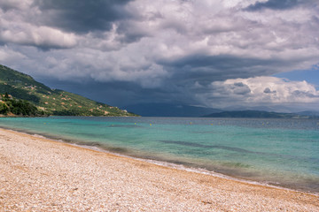 Beautiful landscape – turquoise colored sea water, golden sand, gray sky with dark stormy clouds and mountains on the horizon. 