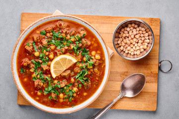 Moroccan Harira Soup in white plate on woden board on grey concrete table top. Harira is Moroccan Cuisine dish with lamb or beef meat, chickpeas, lentils, tomatoes, ciliantro. Ramadan Iftar Food.