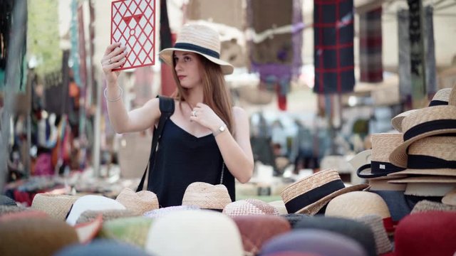 Young woman trying on panama hat at the flea market and looking into the mirror. Summer fashion trends, market trading and beauty concept 