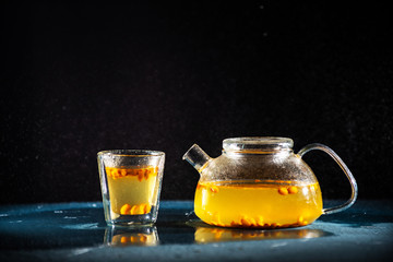 herbal sea buckthorn tea in a transparent teapot and in a transparent glass with a double bottom on a black background with splashes of water