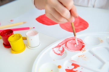 child draws a pink heart in gouache on a white table with a palette