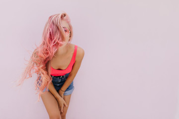 Glamorous young woman with curly pink hairstyle fooling around in studio. Indoor photo of...