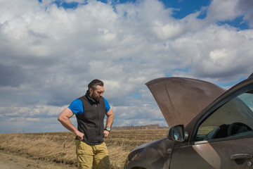 A man near a broken car on a rural road and looks at the engine under the hood.