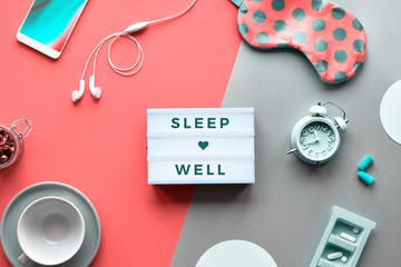 Text "Sleep well" on trendy lightbox. Pills, capsules and calming tea. Flat lay, two tone orange coral and silver background. Coral sleep mask with polka dots, alarm clock, earphones and earplugs.