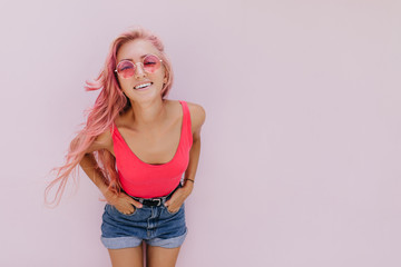 Curly pink-haired girl isolated on white background with hands in pockets. Studio shot of fashionable tanned woman in summer outfit.