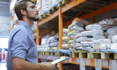 Young man shopping or working in a hardware warehouse standing checking supplies on his tablet.
