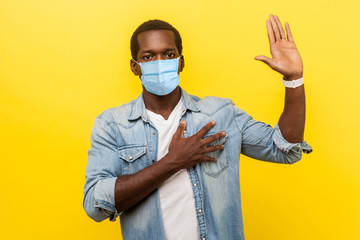 I swear! Portrait of honest serious man in denim casual shirt with surgical medical mask keeping hand on chest, raising palm, giving promise, pledge. indoor studio shot isolated on yellow background