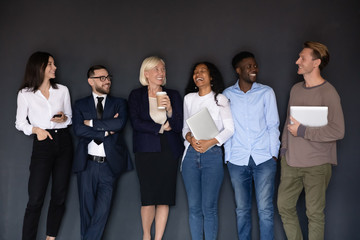 Obraz premium Overjoyed diverse multiethnic businesspeople stand posing near black wall have fun together joking at break, excited happy multiracial colleagues laugh and joke, unity, team spirit concept