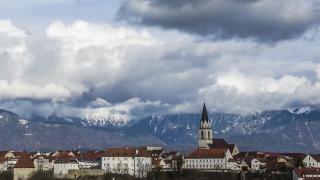 Time lapse zoom in of beautiful sun shinning medieval town Kranj with snow covered mountain range Alps behind.  White clouds form and roll above, moving to the right.