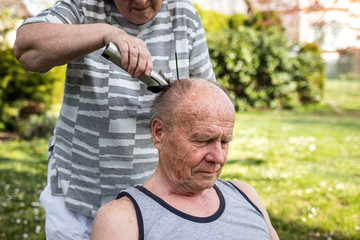 Woman is doing haircut with hair clipper to senior man outdoors at garden. Elderly people take care each other