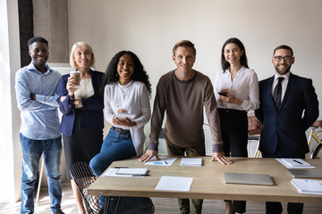 Group portrait of happy multicultural businesspeople stand together look at camera posing at workplace, smiling multiethnic diverse colleagues or employees show unity and support, cooperation concept