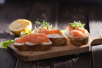 Sandwiches or canapes made of full grain baguette bread, cream cheese and smoked salmon garnished with fresh thyme twigs and leaves of mint and lemon in the background