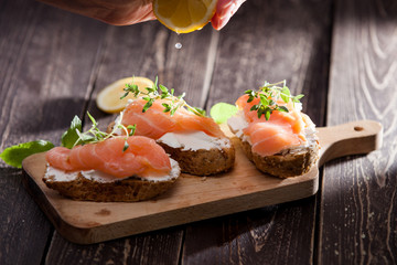 A hand squizeeng lemon juice on sandwiches or canapes made of full grain baguette bread, cream cheese and smoked salmon garnished with fresh thyme twigs and leaves of mint and lemon in the background