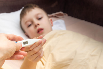 A teenage European boy is lying on the bed. The child is ill. Mom gives her son a thermometer to measure his temperature.
