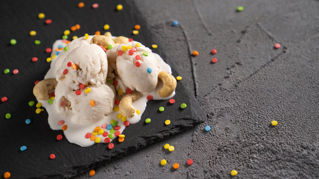 White Ice Cream With Cashew Nuts And Colored Hundreds And Thousands Sprinkles On Black Plate. Melted Vanilla Ice Cream