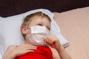 A blond European boy is lying on the bed and coughing.