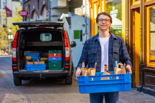 Portret of a happy smiling young entrepreneur delivering wine and food by a local restaurant. With his black transporter car in the background