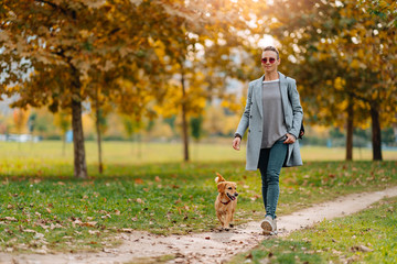 Woman walking on a park trail with a small brown dog in autumn
