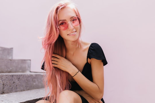 Wonderful caucasian lady sitting on stone stairs and touching her pink hair. Outdoor photo of lovely female model with tanned skin wears black attire.