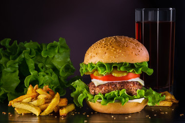 Hamburger with beef cutlet, tomato, pickles, lettuce, and cheese, as well as french fries and a drink in a glass against a dark background