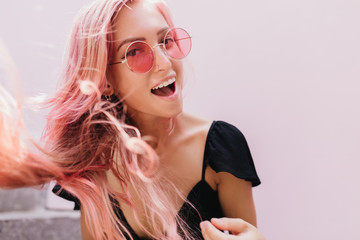 Close-up portrait of inspired pink-haired lady laughing to camera. Photo of ecstatic tanned girl in black dress sitting on stairs.