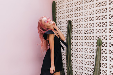 Pretty tanned female model in elegant dress laughing near cactus. Photo of wonderful pink-haired girls wears black clothes in summer day.