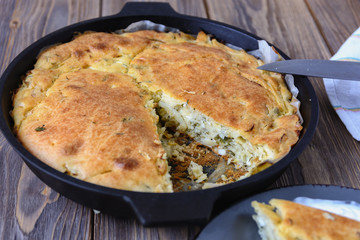 Baked cabbage stuffed pie, ready for dinner in a cast-iron skillet on a wooden board. A piece of cake is cut out for eating.