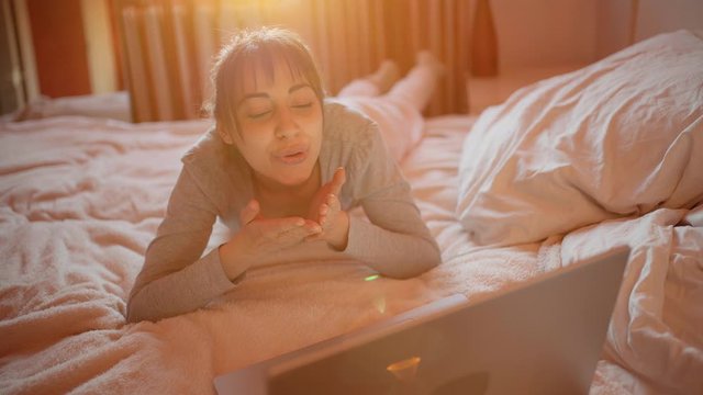 happy excited woman having video chat online, using laptop camera, send air kiss to boyfriend while lying on bed at home. chatting in social network