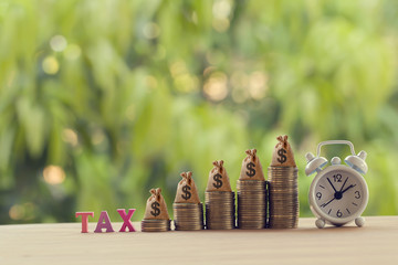 Finance tax concept: Words tax, alarm clock, US money bags on rows of rising coins. depicts ad...